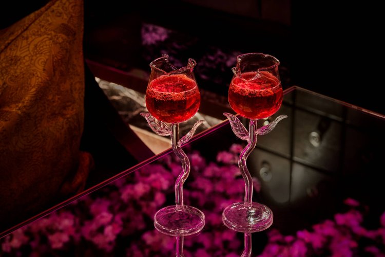 Two elegant rose-shaped glasses filled with a red drink on a reflective surface under pink lighting, perfect for celebrating Mother's Day at Tattu Restaurant.