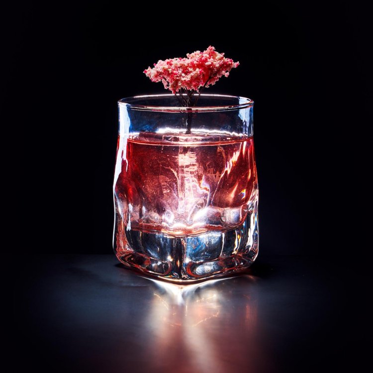A clear glass with a textured base holds a pink liquid, illuminated dramatically against a dark background. A delicate pink flower floats on top, adding an elegant touch to the visually striking composition.