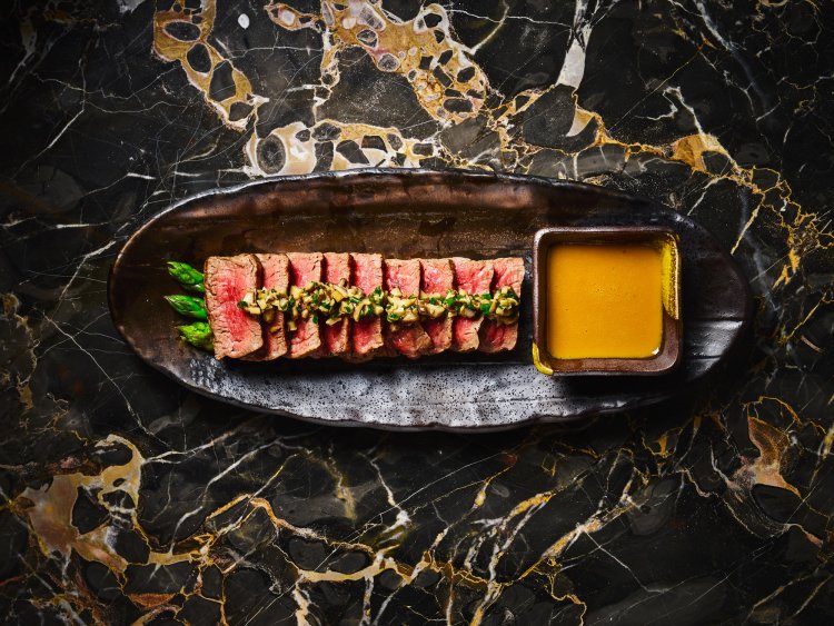 Sliced steak with asparagus and sauce on a dark oval plate, placed on a marbled surface.