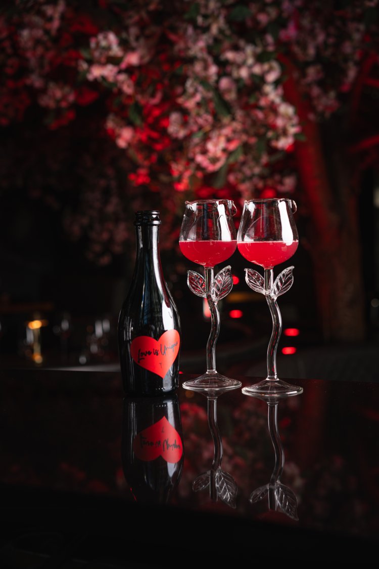 Romantic set-up with rose-themed wine glasses and a wine bottle under cherry blossoms, with red lighting in the background.