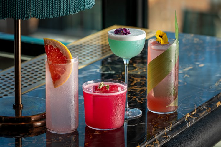 A variety of colorful cocktails sit on a marble bar top, each garnished with flowers, fruit slices, and decorative leaves.