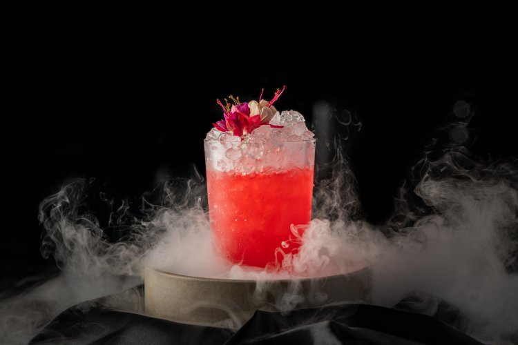 A vibrant red cocktail with ice and edible flowers, surrounded by mist on a round surface.