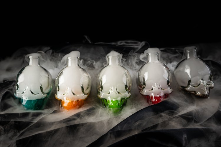 Five skull-shaped bottles with colored bases, emitting smoke, on a dark and misty background.