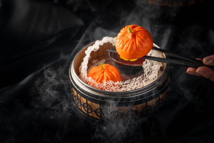 Chopsticks holding a pumpkin-shaped steamed bun over a steaming bamboo basket.