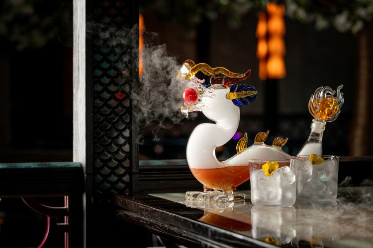 Glass dragon-shaped decanter emitting smoke from its mouth, containing a reddish liquid. Two glasses with ice and lemon peels sit beside it on a dark reflective surface, with blurred warm lighting in the background.
