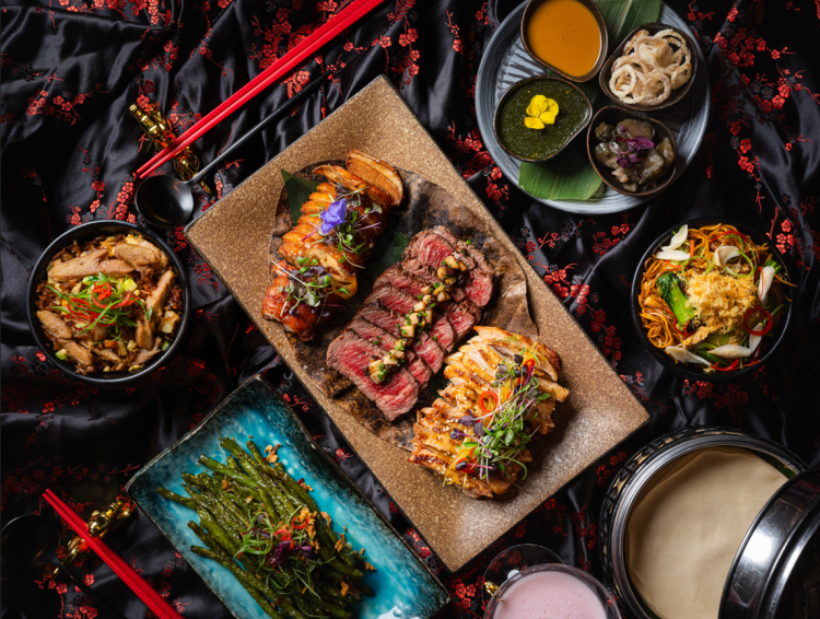 A variety of Asian dishes including sliced meats, vegetables, and garnishes on a patterned tablecloth with chopsticks.