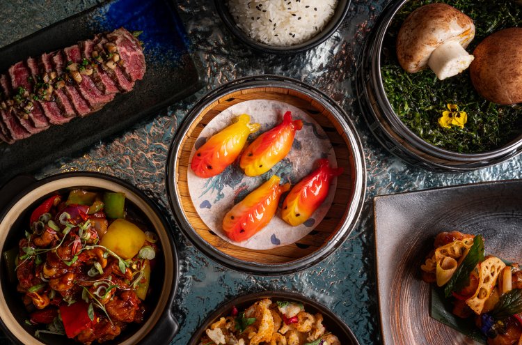An assortment of dishes on a textured surface: sliced steak with garnish, rice, colorful fish-shaped dumplings, mushrooms on herbs, a vegetable stir-fry, crispy fried items, and lotus root garnished with greens.