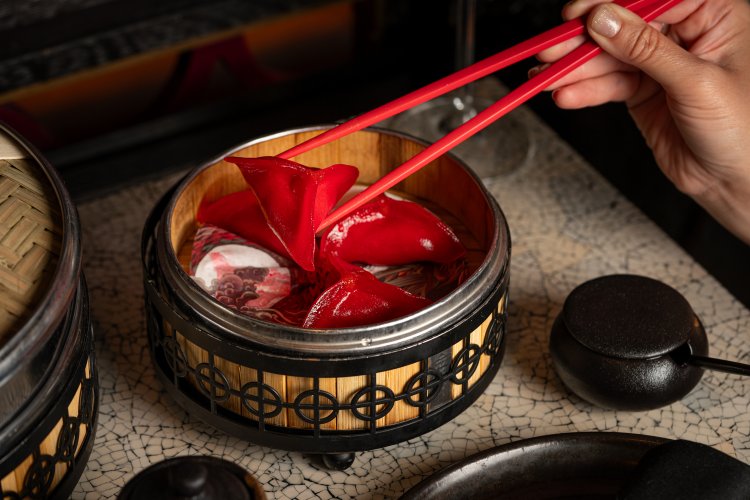 At Tattu Birmingham Restaurant, a person uses red chopsticks to pick up a red dumpling from a decorative bamboo steamer with black accents. The table's mosaic pattern complements the scene, while a dark teapot adds an elegant touch nearby.