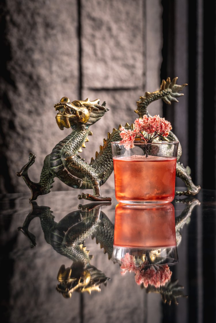 A bronze dragon statue is positioned next to a glass of red cocktail garnished with pink flowers. The setup is on a reflective surface with a stone-like textured background.