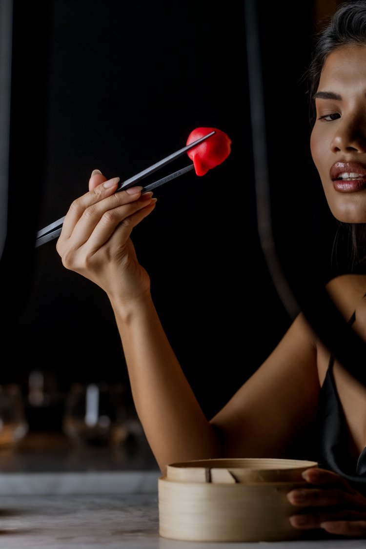 A woman holds a piece of bright red sushi with chopsticks. She is sitting at a table with a bamboo steamer. The background is softly blurred, creating an intimate dining atmosphere.