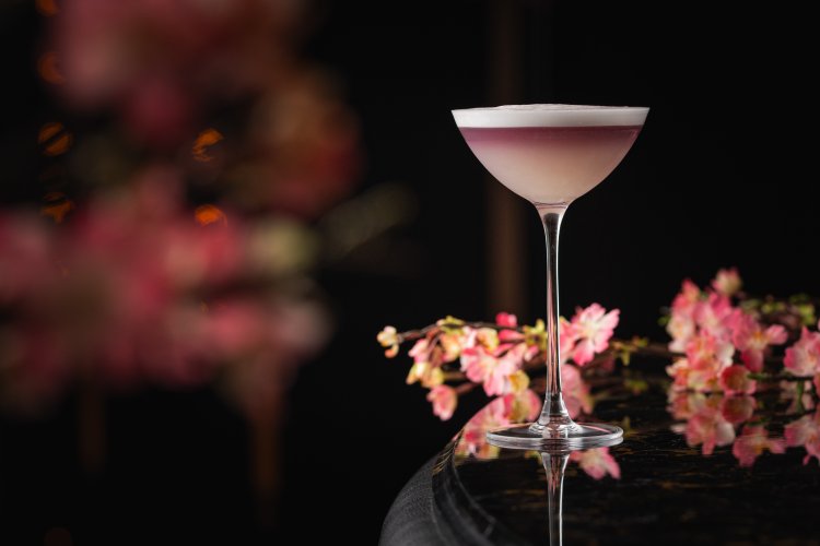 A pink cocktail in a delicate glass rests on a dark, glossy surface. Soft pink flowers surround it, creating a romantic and elegant atmosphere. The blurred background glows with warm light, perfectly capturing the essence of celebrating Mother’s Day in full bloom at Tattu.