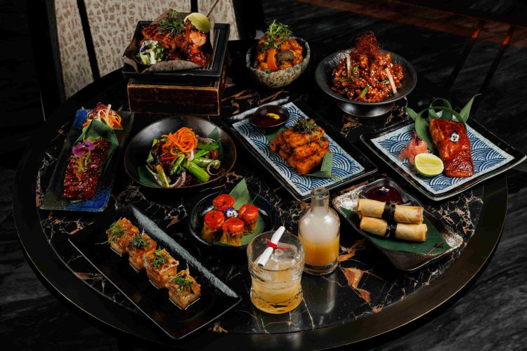 A marble table set with a variety of Asian fusion dishes, including grilled meats, spring rolls, salads, skewers, colorful garnishes, dipping sauces, and two cocktails, all artfully arranged.