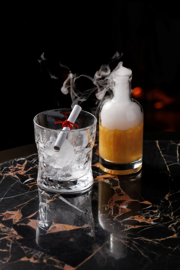 A glass with ice and a rolled paper tied with red ribbon sits next to a small bottle of smoky, brown liquid on a dark marble table—celebrate your graduation with Tattu's "Wisdom of the Dragon" menu for a unique experience.