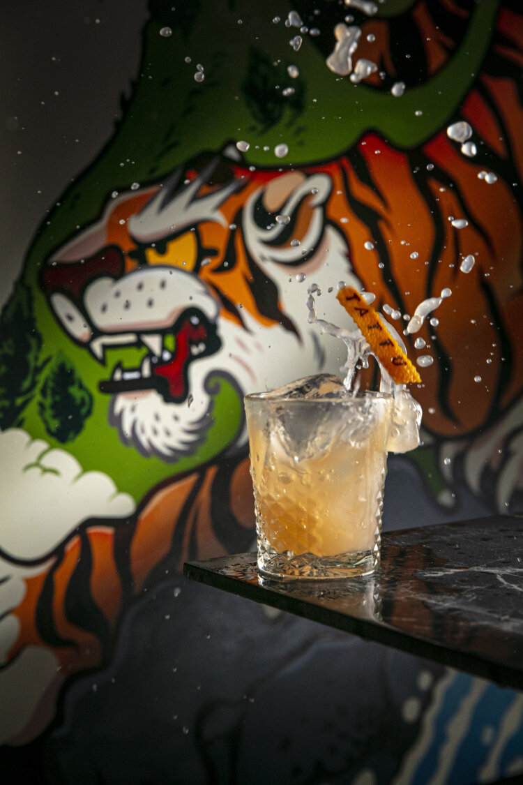 A cocktail splashes in a glass with ice and a dried citrus garnish, set against a vibrant mural of a roaring tiger with orange, white, green, and black colors. Water droplets are frozen mid-air.