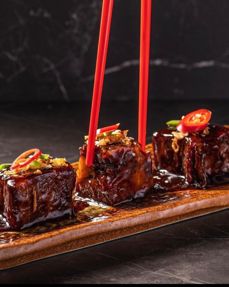 Three glazed, braised pork belly cubes garnished with sliced chili and herbs are arranged on a rectangular ceramic plate. Red chopsticks are lifting one piece. The background is dark and out of focus.