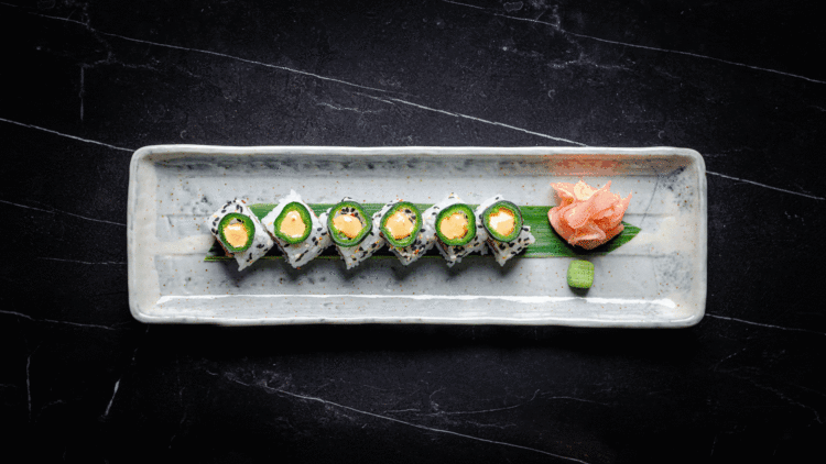 A rectangular ceramic plate with six sushi rolls topped with avocado and sauce, arranged in a row on a green leaf, with pickled ginger and a wasabi ball on the side, set against a dark background.