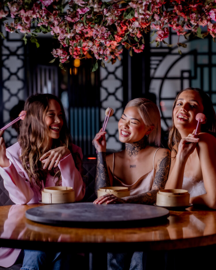 Three women sit at a round table with bamboo steamers, smiling and holding pink chopsticks under hanging pink flowers in a restaurant with decorative screens—Celebrate Your Special Occasion in Style at Tattu Restaurants.